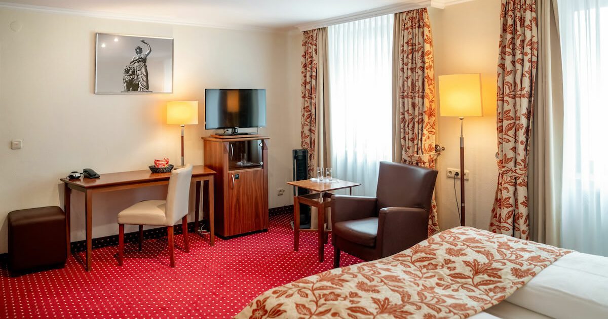Hotel_Europaeischer_Hof_Doppel_Komfort_Innenhof_Hotelzimmer_3 Ausschnitt aus einem günstigen Doppelzimmer in München mit Schreibtisch, Stuhl, Tischlampe, Fernseher, einem Sessel an einem Tisch, einem Bett und rot/weißen Vorhängen