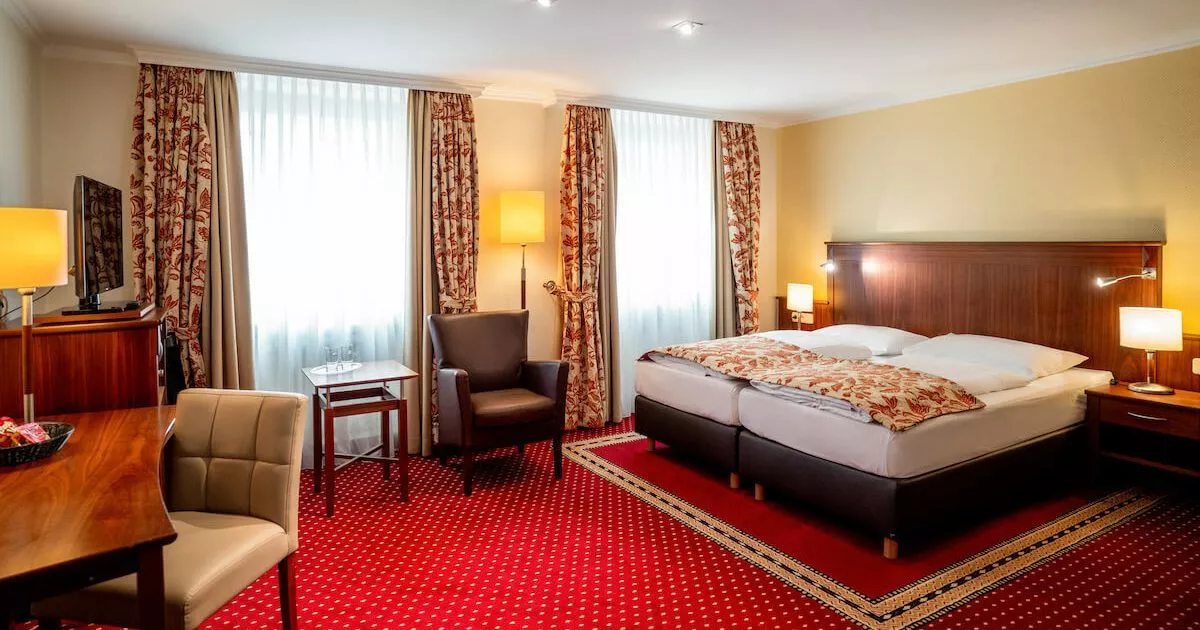 Ein voll ausgestattetes Doppelzimmer mit Bett, Sessel, TV, Schreibtisch und Stuhl