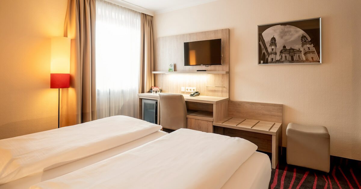 Hotel_Europaeischer_Hof_Doppel_Standard_City_Hotelzimmer_1 Schreibtisch mit Stuhl, Kofferablage, Fernseher, Hocker sowie Teil eines Bettes in einem günstigen Doppelzimmer in München