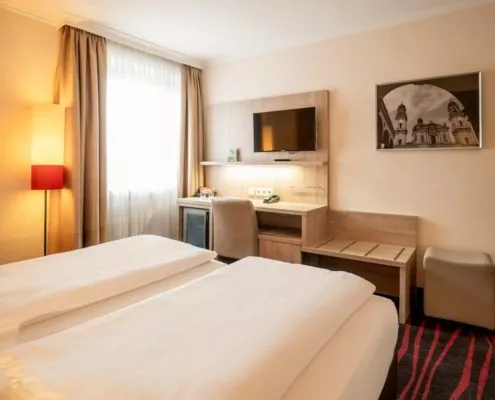 Schreibtisch mit Stuhl, Kofferablage, Fernseher, Hocker sowie Teil eines Bettes in einem günstigen Doppelzimmer in München