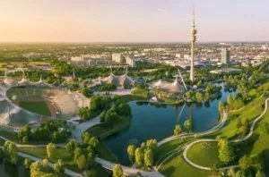 Olympiaturm, den Olympiasee und den grünen Olympiaberg an einem Sommerabend. Im Hintergrund ist die Stadt München zu sehen