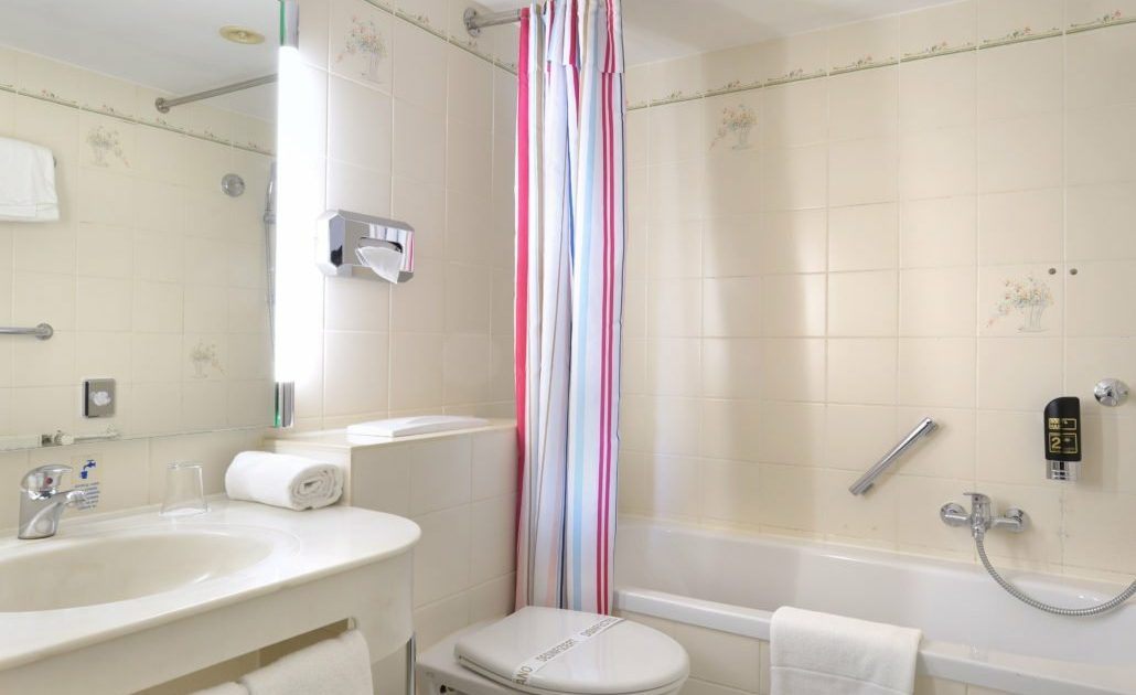 DZ_und_Schlafcouch05-1030x771 Badezimmer mit Badewanne, buntem Duschvorhang, Toilette und Waschbecken mit Spiegel in einem günstigen Dreibettzimmer in München