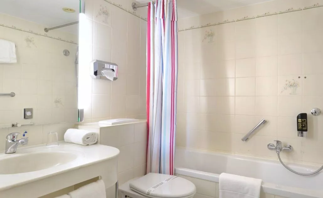 Badezimmer mit Badewanne, buntem Duschvorhang, Toilette und Waschbecken mit Spiegel in einem günstigen Dreibettzimmer in München