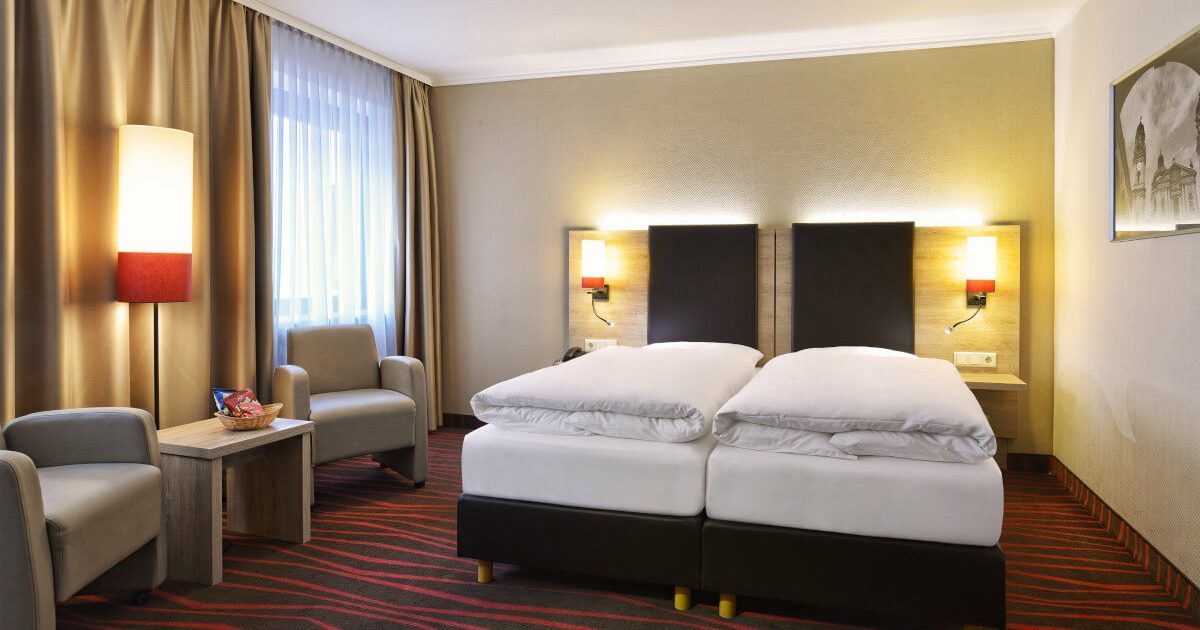 Doppel-Zweibett Innenhof Standard Helles, modernes und günstiges Doppelzimmer in München mit Bett, zwei grauen Sesseln und beigen Vorhängen