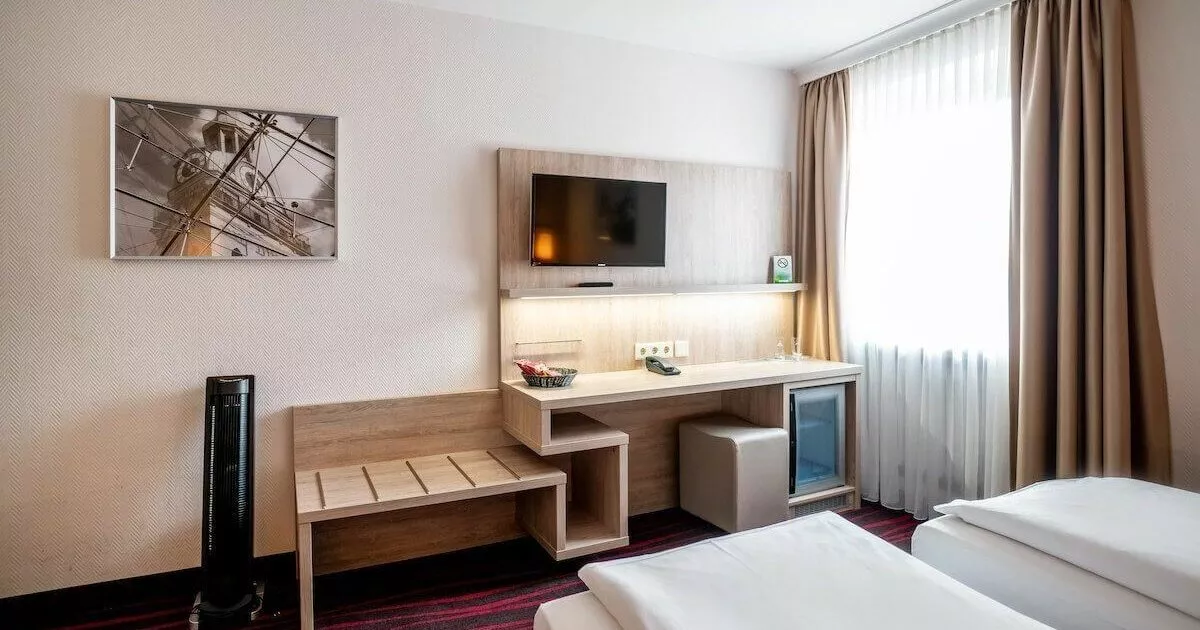 Ausschnitt aus einem günstigen Doppelzimmer in München mit Schreibtisch, Hocker, Fernseher, einem Bett und beigen Vorhänge