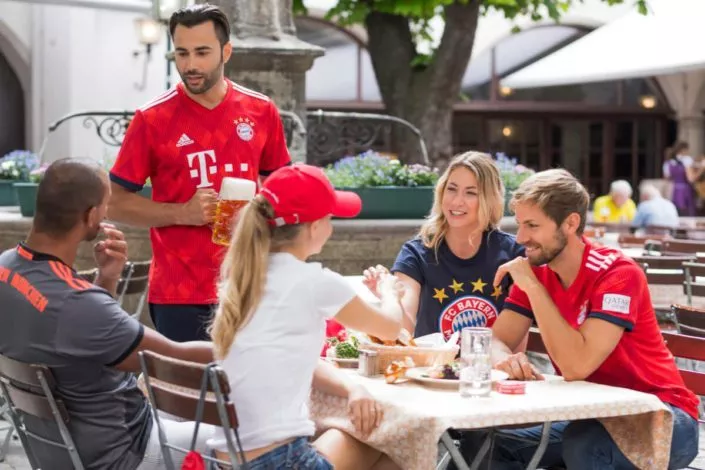 Fußballfans im FC Bayern München Trikot, welche an einem Tisch mit Essen und Bier im Freien sitzen