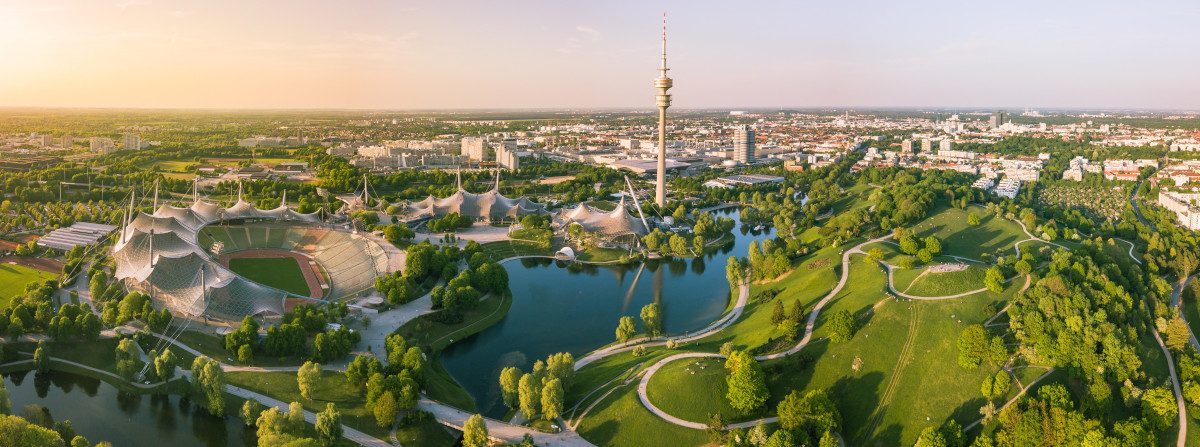 Olympiapark in München mit Blick auf das Olympiastadion, die Olympiahalle, den Olympiaturm, den Olympiasee und den grünen Olympiaberg an einem Sommerabend. Im Hintergrund ist die Stadt München zu sehen