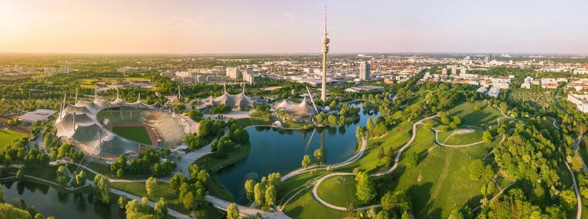 Blick von oben auf den Olympiapark in München mit Blich auf das Olympiastadion, die Olympiahalle, den Olympiaturm, den Olympiasee und den grünen Olympiaberg an einem Sommerabend. Im Hintergrund ist die Stadt München zu sehen