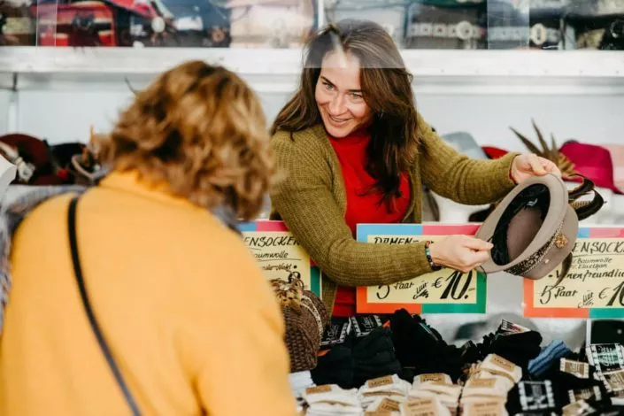 Verkäuferin redet mit einer Kundin an einem Verkaufsstand für Hüte, Socken, Taschen und Ähnliches