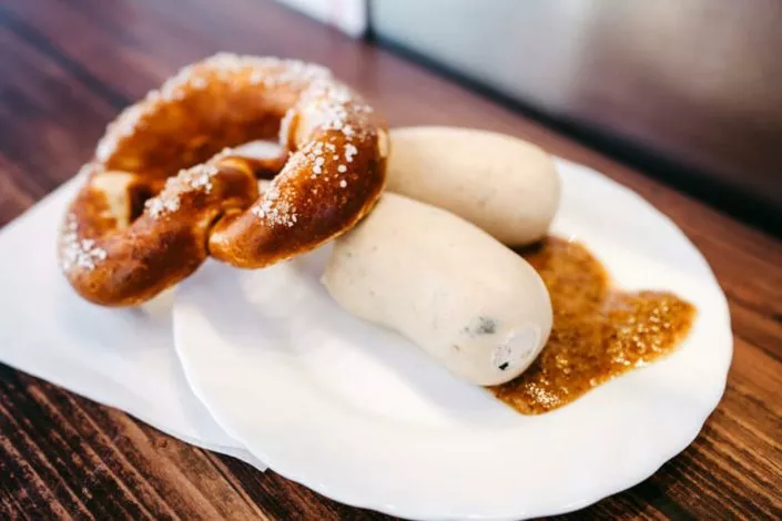 Teller mit zwei Weißwürsten, süßem Senf und einer Brezel auf einem Holztisch